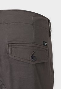 Tumši pelēkas bikses ar aizmugurē poga aiztaisāmu kabatu, gludu tekstūru un smalkām šuvju detaļām. Kabatā redzama zīmola etiķete.