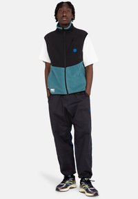 Fleece vest in black and teal, zippered front with chest pocket, paired with dark pants featuring side accents and multicolored sneakers.