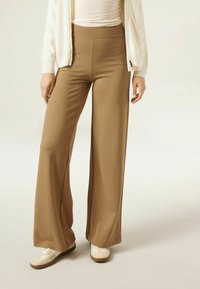 Pantalons larges en tissu marron clair, avec une taille haute, une texture lisse et une coupe légèrement évasée. Associés à des baskets crème.