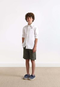 Witte button-up shirt, groene shorts met blauwe patronen, grijze schoenen. Jongen staat rechtop tegen een eenvoudige achtergrond.