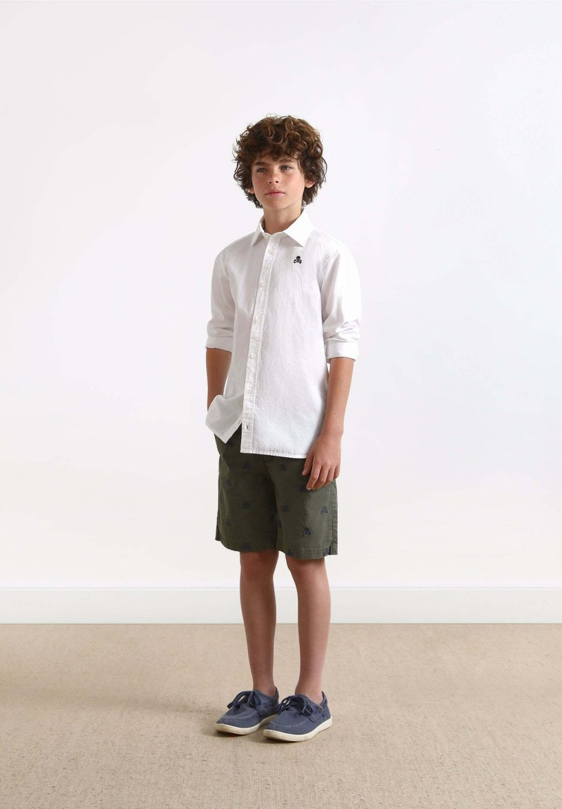Witte button-up shirt, groene shorts met blauwe patronen, grijze schoenen. Jongen staat rechtop tegen een eenvoudige achtergrond.