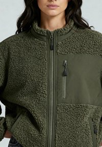Veste polaire vert olive à finition texturée et duveteuse. Dispose de poches zippées, d'un col montant et de panneaux contrastants en tissu lisse.