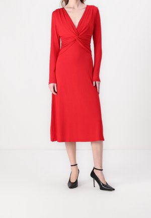 Femme portant une robe midi rouge à manches longues avec un détail torsadé à l'avant, chaussée de talons hauts noirs à bride cheville, se tenant devant un fond blanc.