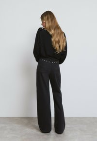 Un pull noir avec des boutons décoratifs dans le dos, associé à un pantalon large noir taille haute avec des accents cloutés sur la ceinture.