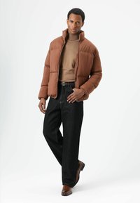 Bruine donsjas met een hoge kraag, gedragen over een beige coltrui, gecombineerd met donkere denim broek en bruine leren schoenen.