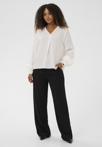 Blouse blanche à manches longues avec un col en V et des poignets froncés, associée à un pantalon noir à jambes larges pour une silhouette équilibrée.