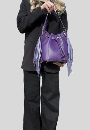 Persona in abito nero che tiene una borsa secchiello in pelle viola con occhielli d'argento e lunghe frange su entrambi i lati.