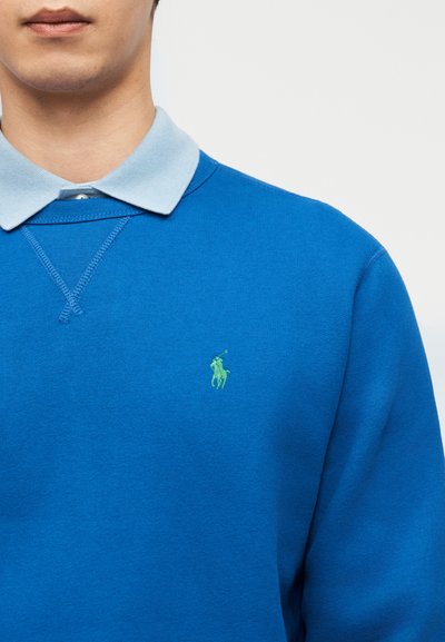 Άνδρας που φοράει μπλε φούτερ πάνω από ανοιχτό γαλάζιο πουκάμισο με γιακά, προβάλλοντας το μικρό πράσινο λογότυπο παίκτη πόλο στο στήθος του φούτερ.