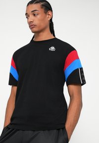 Tricou negru cu panouri roșii și albastre pe umeri, având un mic logo alb Kappa pe piept. Mâneci scurte, material din bumbac.