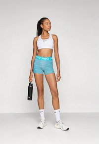 Nike Pro sportinė liemenėlė balta spalva, derinama su aqua mėlynomis šortais, kurių juostos juosta kontrastinga. Modelis laiko juodą vandens butelį, avėdamas baltus sportbačius.