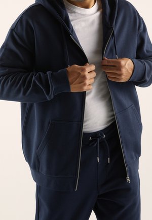 Donker marineblauwe hoodie met rits, twee voorkruiszakken en geribde manchetten. Draagt over een wit T-shirt, gecombineerd met bijpassende broek.