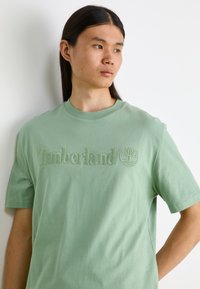Ανοιχτό πράσινο βαμβακερό μπλουζάκι με ανάγλυφο λογότυπο "Timberland" και έμβλημα δέντρου. Κλασική στρογγυλή λαιμόκοψη και κοντά μανίκια με λεία υφή.