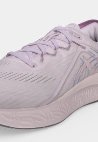 Gros plan d’une chaussure de course rose clair avec des lacets blancs, un subtil motif brodé sur le côté et une semelle texturée pour l’adhérence.