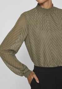 Femme portant un chemisier transparent vert olive avec un motif en zigzag et un col smocké, associé à un pantalon noir taille haute, la main dans la poche.