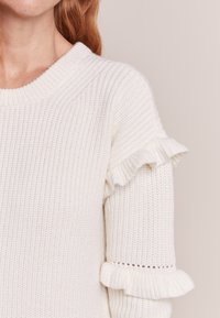 Biała, dziana sweter o prążkowanej fakturze, okrągłym dekolcie i falbaniastych zdobieniach przy rękawach. Materiał wygląda na miękki i lekki.