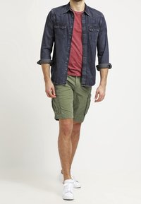 Tmavě modrá denimová košile s knoflíky přes červené tričko, doplněná zelenými cargo šortkami a bílými teniskami. Obsahuje dvě náprsní kapsy.