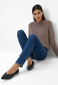 Maglione marrone a coste, jeans blu slim-fit e scarpe nere di pelle piatte. Modello seduto con le gambe incrociate, sfondo minimale.