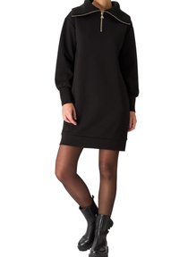 Femme portant une mini-robe noire à manches longues avec un grand col zippé et des bottines noires, debout les jambes croisées.