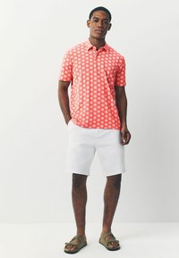 Camisa polo de manga corta para hombre en color coral con un patrón de explosión de estrellas en blanco, combinada con pantalones cortos blancos y sandalias marrones.