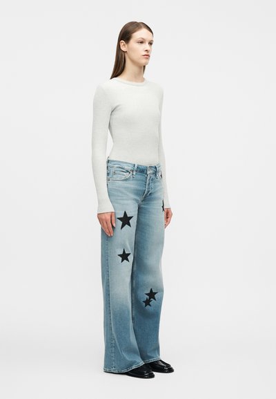 Γυναίκα που στέκεται, φοράει ανοιχτό γκρι μακρυμάνικη μπλούζα, πλατειά μπλε τζιν με μαύρα αστέρια και μαύρα παπούτσια, με φόντο λευκό.