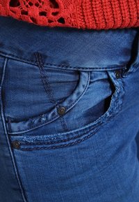 Jean en denim bleu avec une poche avant munie de boutons noirs et un détail cousu ; le tissu a une texture lisse et un léger effet délavé.