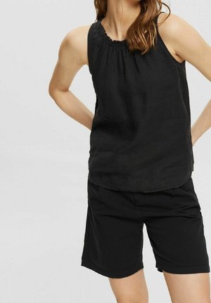 Vrouw draagt een mouwloze zwarte top met een geplooide halslijn en zwarte knielange shorts, handen rusten op de heupen tegen een effen achtergrond.