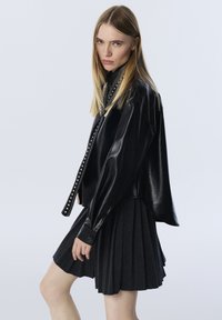 Giubbotto di pelle nera con vestibilità comoda e tondini d'argento sporgenti lungo il davanti, abbinato a una gonna plissettata di un grigio scuro.