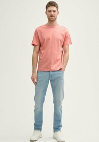 Uomo in piedi con una mano in tasca, indossando una maglietta a maniche corte color corallo, jeans azzurri chiari e sneakers bianche su uno sfondo semplice.