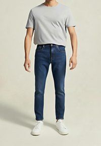 Homme portant un t-shirt gris clair, un jean slim bleu foncé et des baskets blanches, debout devant un fond uni.