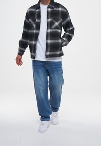 Person klädd i en svart och vit rutig jacka, vit t-shirt, löst sittande blå jeans och vita sneakers som går mot en vit bakgrund.