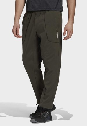 Man die donkergroene Adidas Terrex-broek en zwarte outdoor wandelschoenen draagt, staand tegen een effen achtergrond.