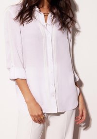 Femme portant un chemisier blanc à manches longues boutonné avec une bordure en dentelle et un pantalon blanc, accessoirisée de bagues en or et d'un collier à plusieurs couches.
