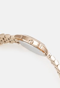 Montre en métal doré avec un boîtier rectangulaire, lunette lisse et bracelet texturé composé de segments liés. Verre transparent en haut.