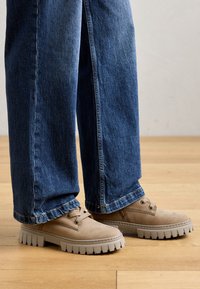 Chaussures en suède beige avec une semelle épaisse et striée grise, assorties à un jean en denim bleu à jambes larges. Les chaussures présentent des lacets ronds et des accents de couture.