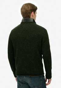 Suéter de punto verde oscuro con un acabado texturizado, que presenta un cuello doblado con un forro de cuadros, usado sobre unos jeans de mezclilla.