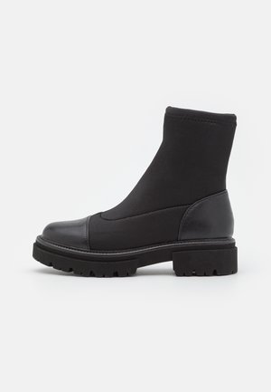 Schwarzer Knöchelstiefel mit klobiger Sohle, elastischem Obermaterial und Lederzehenkappe sowie Fersenpartie, seitlich auf weißem Hintergrund dargestellt.