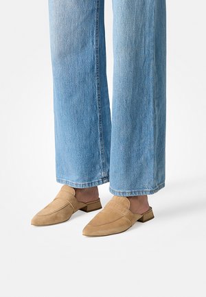 Persoon draagt lichtblauwe wijde spijkerbroek en lichtbruine suède mocassins zonder achterkant, staand op een witte vloer.