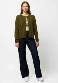 Olivgrön kofta med knappstängning över en krämfärgad randig skjorta, ihopsatt med mörkblå utsvängda jeans och vita sneakers.