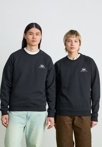 Melni crewneck sporta krekli ar raglāna piedurknēm ir ar baltu logo izšuvumu krūšu apvidū. Modeļi valkā kontrastējošas bikses: gaišo džinsu un rakstainas.