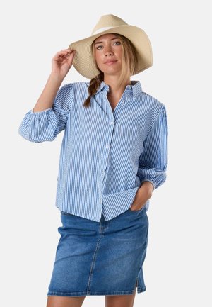 Frau mit geflochtenem Haar trägt einen breitkrempigen Hut, ein blau-weiß gestreiftes Hemd mit Knöpfen und einen knielangen Jeansrock und posiert mit einer Hand in der Tasche.
