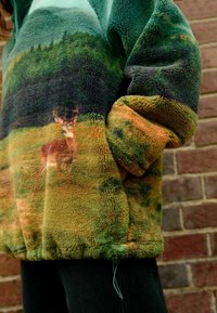 Pull en polaire avec un motif de paysage abstrait vert et marron, présentant une image de cerf. Tissu doux et texturé avec un ourlet à cordon de serrage.