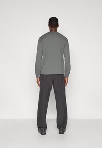 Donkergrijs shirt met lange mouwen en een rechte zoom, gecombineerd met zwarte, loszittende broek en zwarte schoenen, gezien vanaf de achterkant.