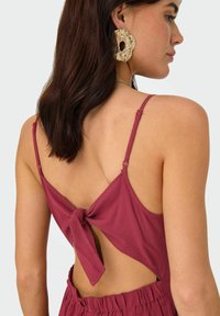 Robe bordeaux avec des bretelles fines, dos nu et détail noué. Présente des boucles d'oreilles texturées en or, avec les cheveux lisses tombant sur une épaule.