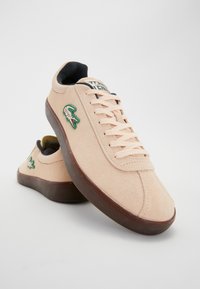 Baskets en daim beige avec semelles marron, design à lacets et logo crocodile vert brodé sur le côté, présentées sur fond blanc.