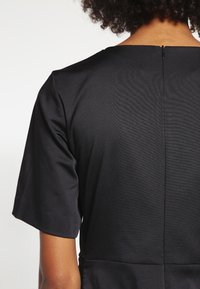 Černé šaty s hladkou texturou, krátkými rukávy a uzavřením na klíčovou dírku na zádech. Design se vyznačuje čistými liniemi a přiléhavým střihem.