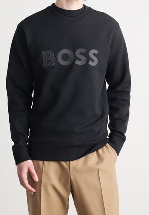 Schwarzer Sweatshirt aus Baumwollmix mit Rundhalsausschnitt, mit einem erhöhten "BOSS"-Logo in einem leicht helleren Farbton. Kombiniert mit beigen Hosen.