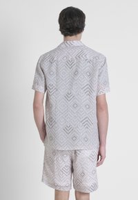 Uomo che indossa una camicia a maniche corte e pantaloncini abbinati beige con un motivo geometrico a diamante, visto di spalle contro uno sfondo neutro.