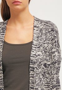 Cardigan en tricot avec un motif texturé crème et marine, design ouvert à l'avant et bords côtelés. Superposé sur un débardeur sombre.