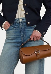 Brun lærveske med klaffelukking, sølvfarget hardware og avtakbar stropp. Kombinert med en mørk jakke og lyse denimjeans.