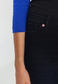 Mörkblå skinny jeans med en slät textur, med en kopparknapp och subtila sömdetaljer. En blå långärmad topp som delvis är synlig.